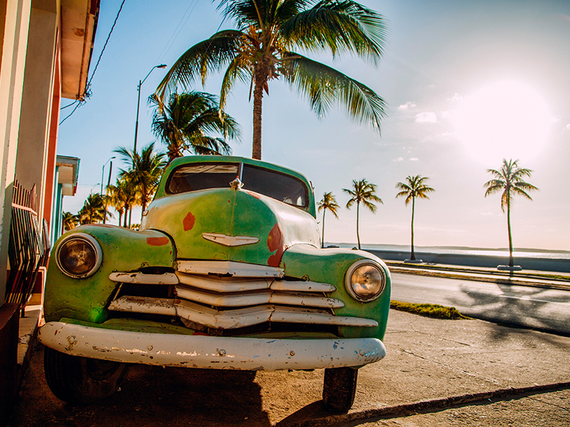 Oldtimer mit Palmen und Meer im Hintergrund.