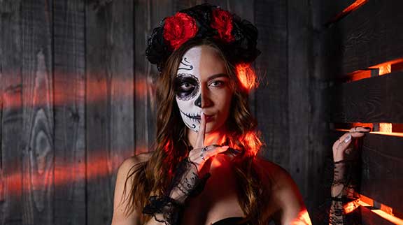 Frau mit halbem Totenkopf-Make-up und rotem Blumenkranz im Haar, inspiriert vom Día de los Muertos, hält den Finger vor den Mund