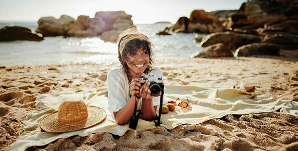 Frau liegt auf einer Decke am Strand bei Sonnenschein und hält lächelnd eine Kamera in der Hand.