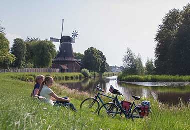 Zwei Radfahrer sitzen an einem Fluss, im Hintergrund ist eine Windmühle.