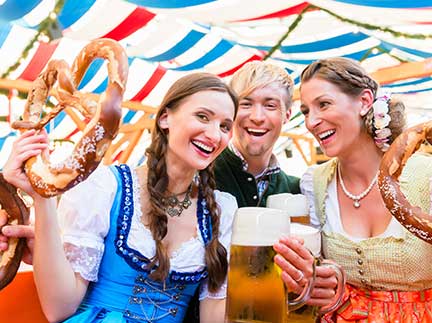 Drei Besucher des Oktoberfestes in Trachten im Zelt mit Bier und Brezeln.
