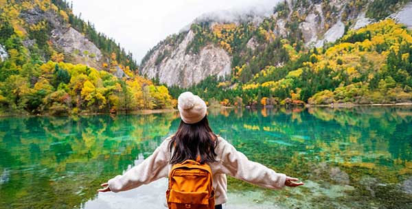 Frau mit Mütze steht vor einen türkisfarbenen See, im Hintergrund sind Berge und bunte Blätter an Bäumen.