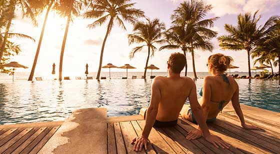 Paar sitzt am Pool bei Sonnenuntergang und schaut auf Meer und Palmen.