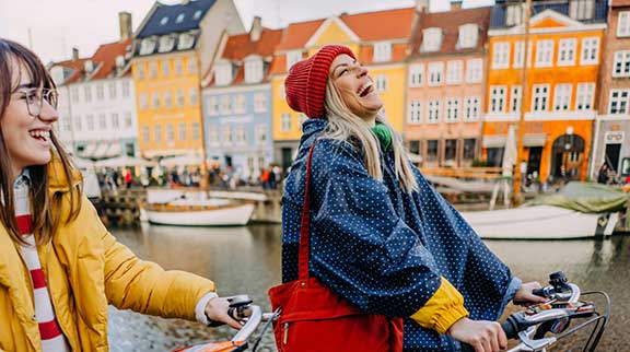 Zwei Frauen fahren herbstlich angezogen mit dem Rad am Hafen einer Stadt mit Bunten Häusern und lachen.